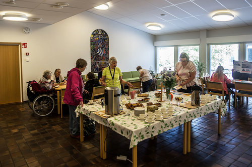 Karhulan seurakuntakeskuksen kahvio. Pöytään on katettu kahvit ja pullat, pöydän äärellä ihmisiä.