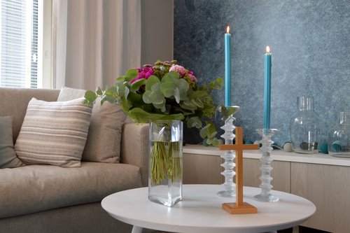 Sohva ja sohvapöydällä kukkia maljakossa sekä kynttilöitä ja risti.