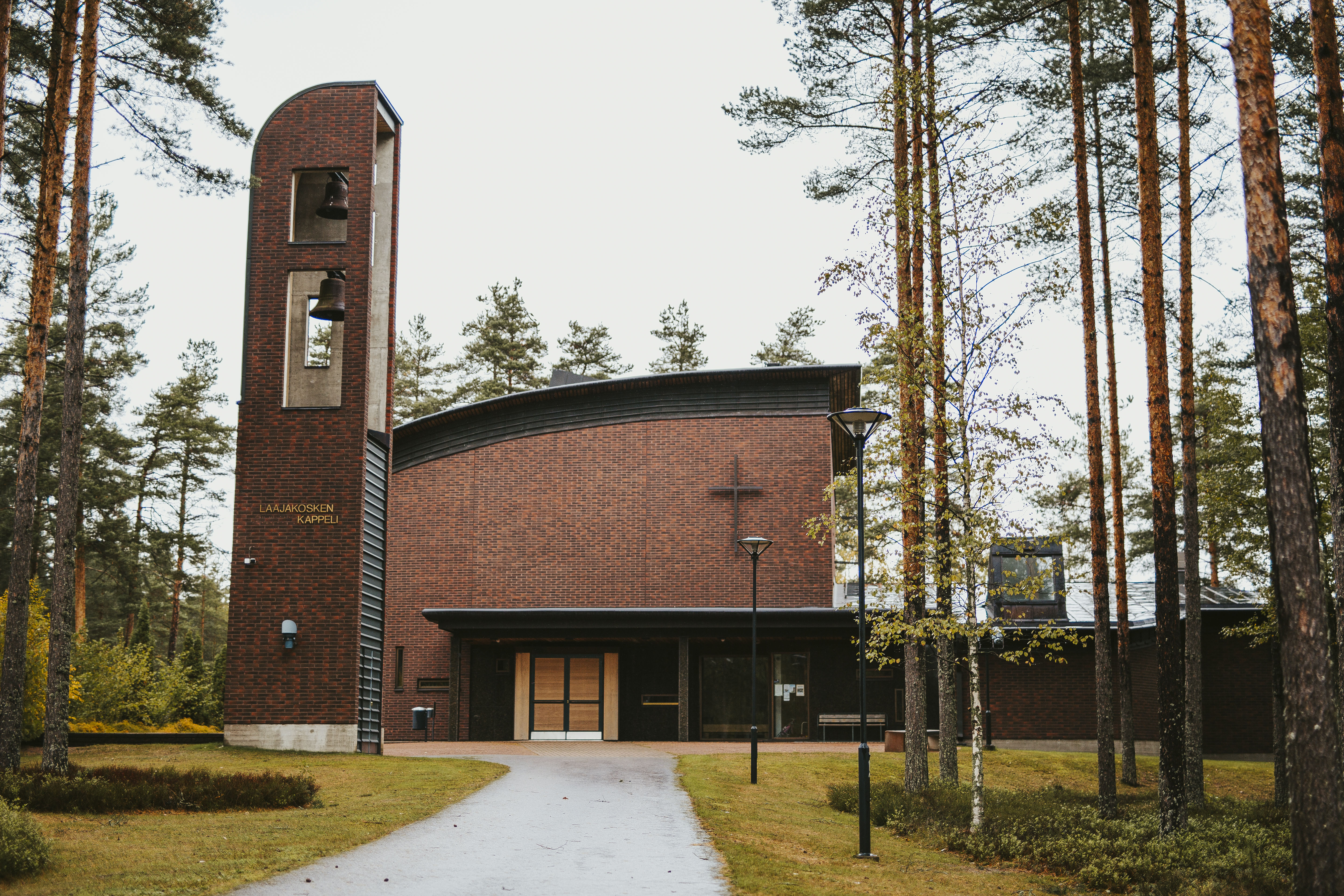 Ruskeatiilinen kappelirakennus ja kellotorni asfalttitien päässä. Tien reunalla puita.