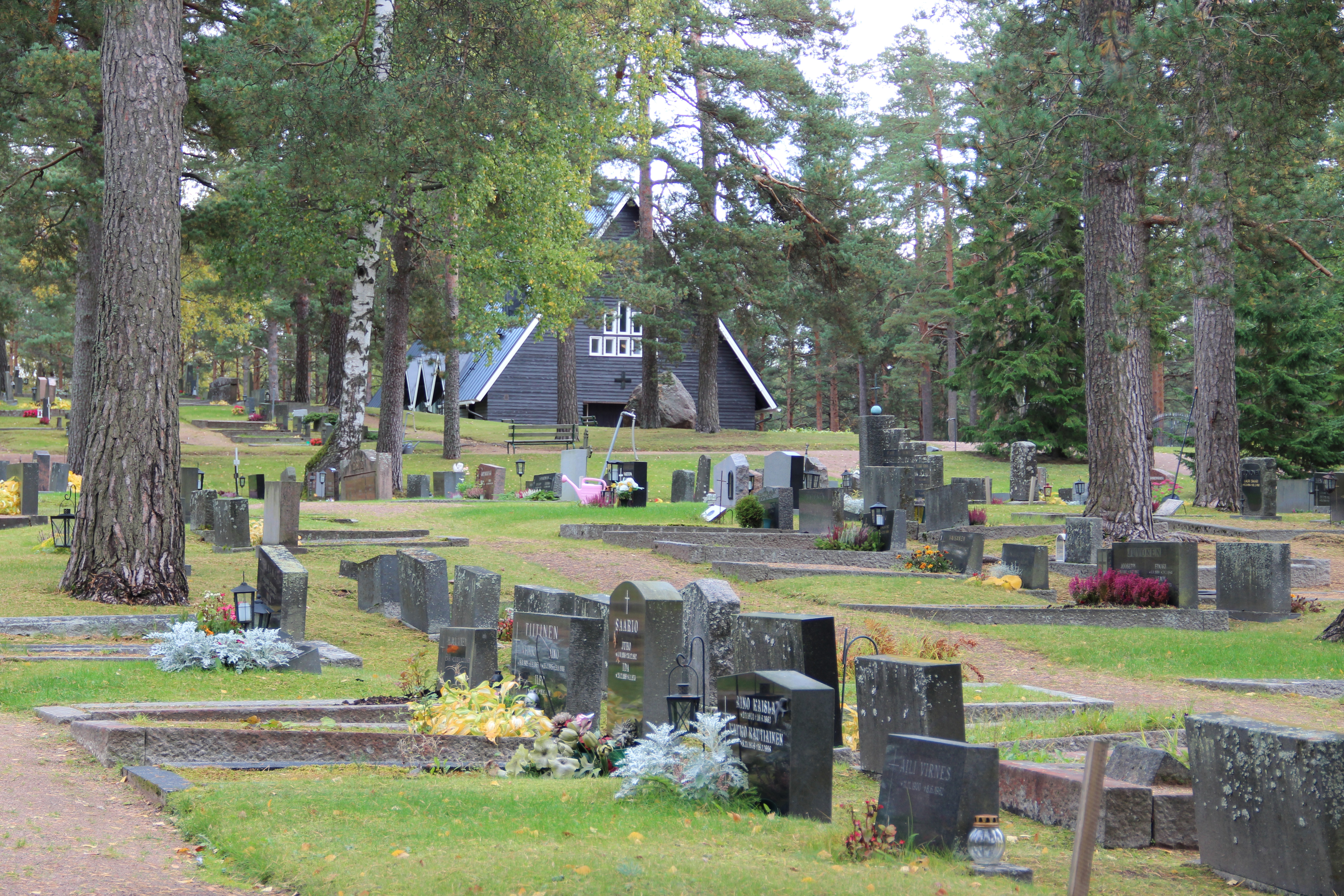 Etualalla paljon hautakiviä, taustalla näkyy tummanharmaa, kolmionmuotoinen kappelirakennus.