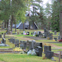 Metsäkulman hautausmaa Etualalla paljon hautakiviä, taustalla näkyy tummanharmaa, kolmionmuotoinen kappelirakennus.