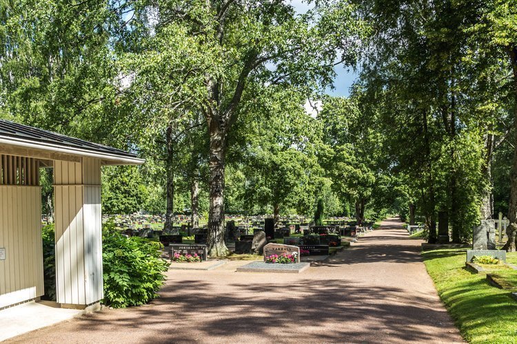 Yleiskuva kesäisestä hautausmaasta, etualalla huoltorakennus ja hiekkatietä.