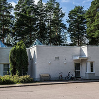 Mussalon seurakuntakoti Valkoinen tiilirakennus, edessä asfalttikenttä, takana metsää.