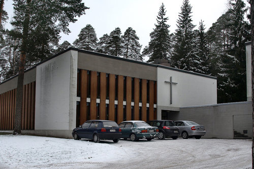 Sunilan seurakuntatalo Valkoinen rakennus, jonka julkisivussa myös ruskeita puupalkkeja. Edessä autoja ja luminen piha.