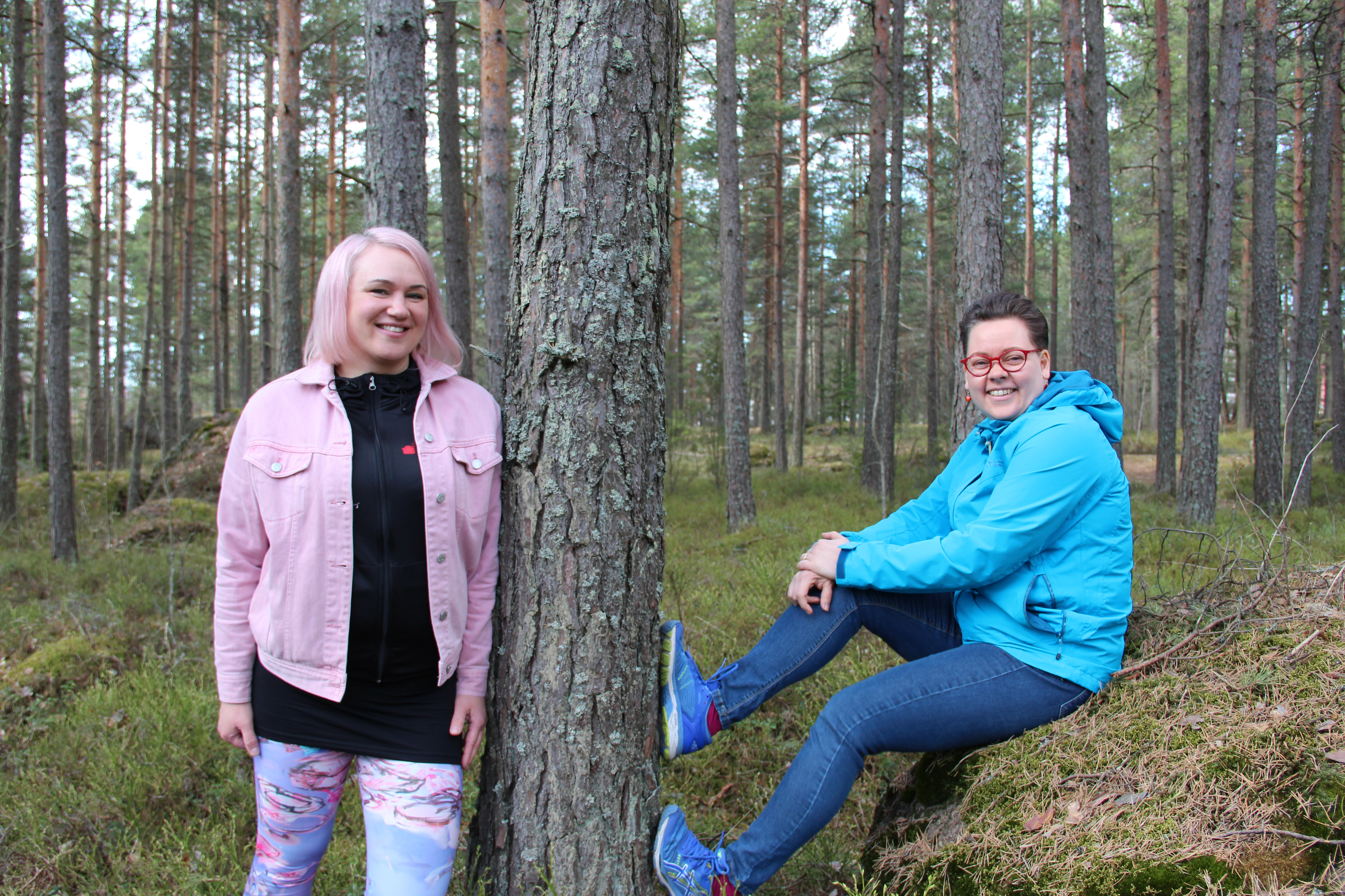 Kaksi naista metsässä, toinen istuu kivellä, toinen nojaa puun runkoon.