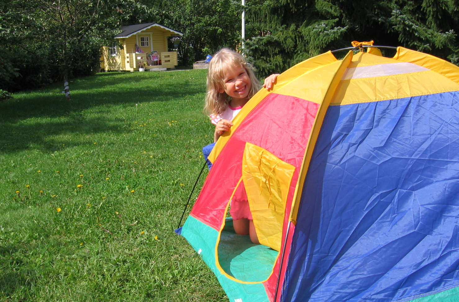 Pieni tyttö kurkistaa värikkään lasten teltan takaa. Pihapiiri, taustalla leikkimökki.