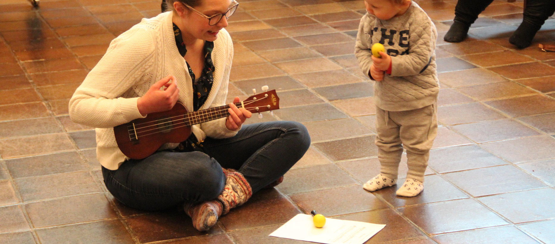 Nainen istuu lattialla ja soittaa ukuleleä, pieni lapsi katsoo vieressä.