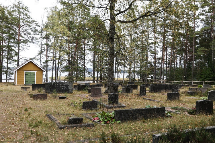 Yleiskuva hautausmaalta, hautakiviä ja hiekkapolkuja. Taustalla huoltorakennus ja meri.