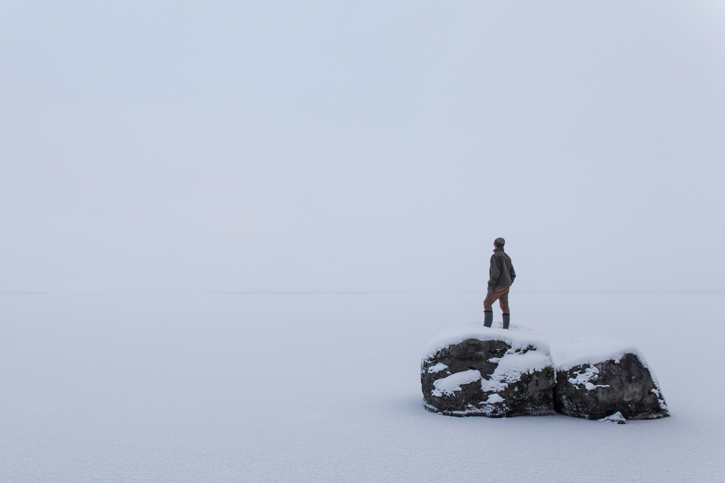 Mies seisoo kivellä ja katsoo meren jäälle - jää ja taivas ovat yhtä harmaat.