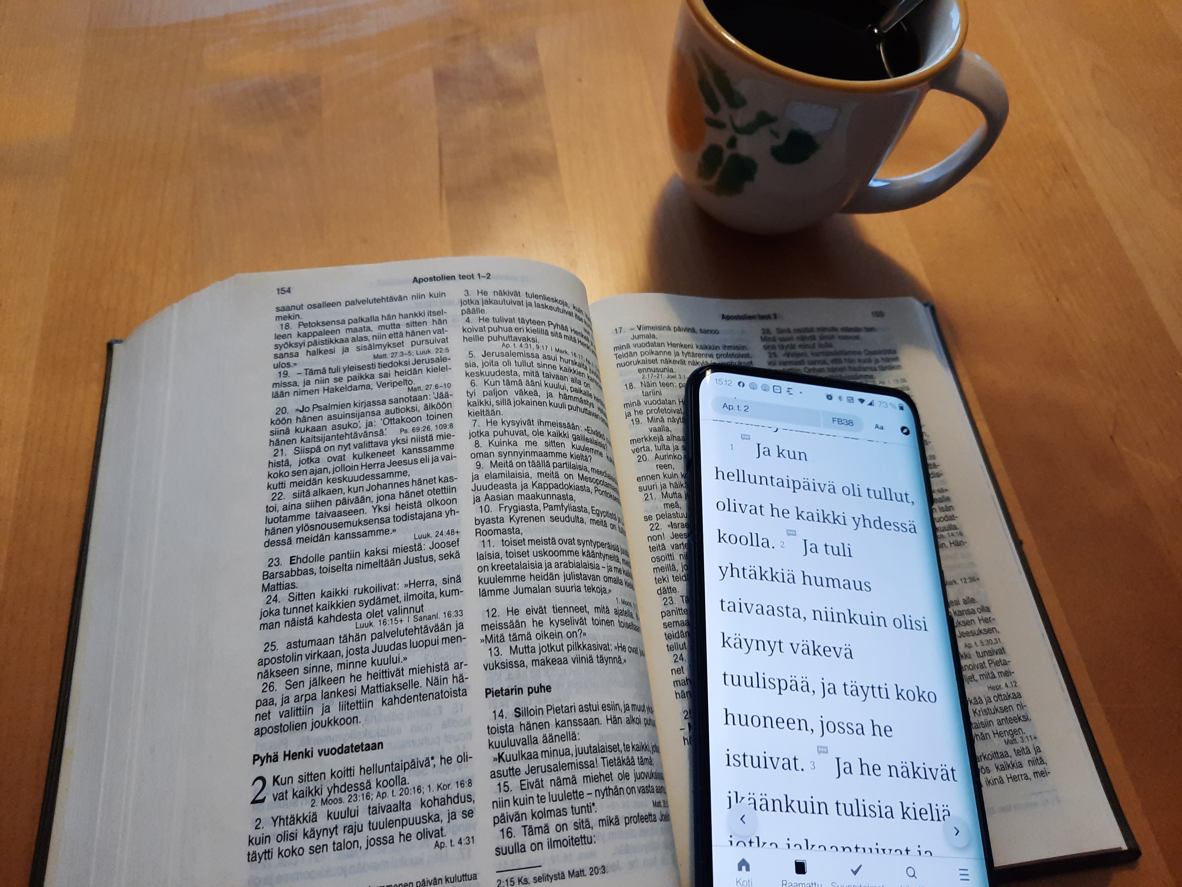 Raamattu pöydällä. Raamatun päällä kännykkä jossa avattuna Raamattusovellus. Vieressä kahvikuppi.