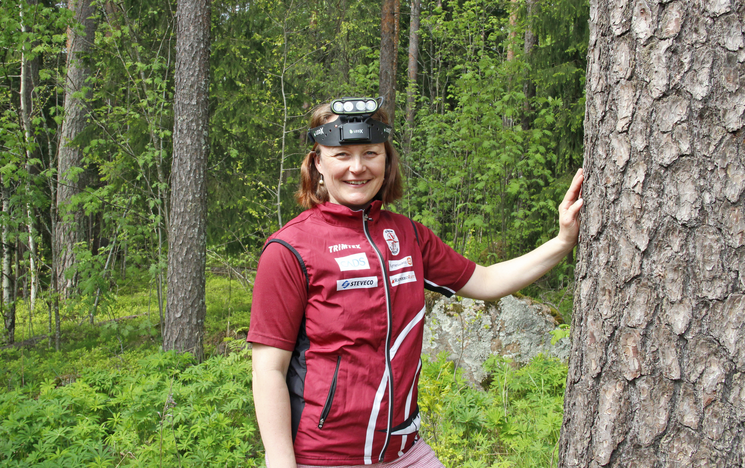 Nainen suunnistusvaatteissa nojaa puunrunkoon.