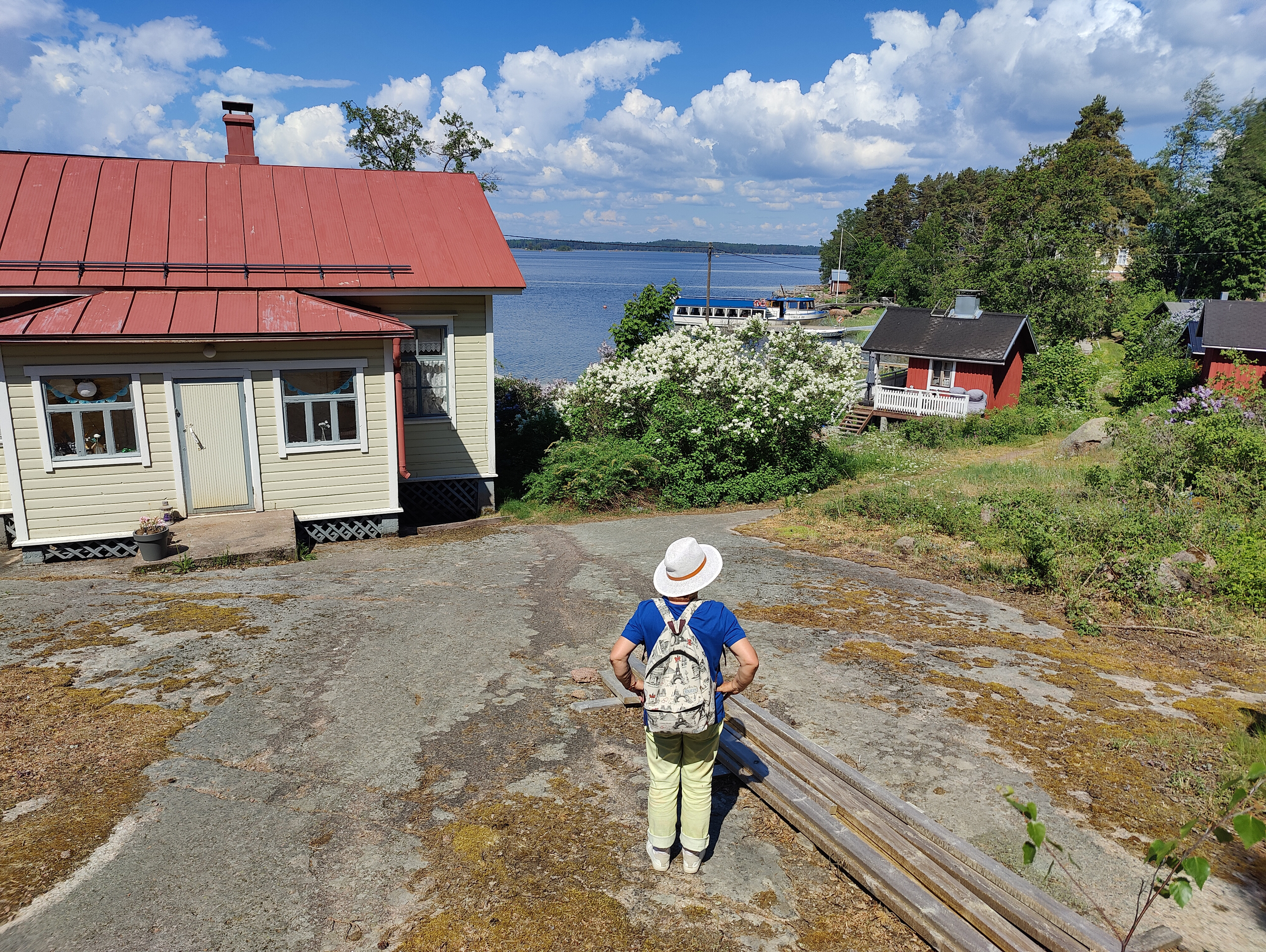 Saaristomaisema, vanha puutalo ja meri taustalla, edessä lierihattuinen ihminen selkä kameraan päin.
